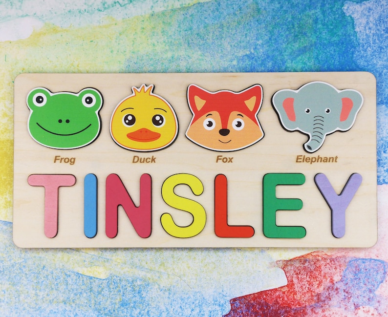 May include: A wooden personalized puzzle with the name "TINSLEY" spelled out in colorful letters. The puzzle also features four animal faces: a green frog, a yellow duck, an orange fox, and a gray elephant.