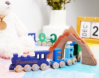May include: A wooden toy train set featuring a dark blue engine with the name Francesca engraved on the side, followed by light blue letter cars spelling ER, passing through a layered wooden mountain tunnel with small wheels on each piece.