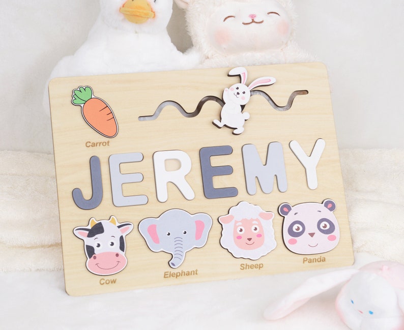 May include: A wooden personalised puzzle with the name "JEREMY" in grey letters. The puzzle features a cow, elephant, sheep, panda, rabbit, and carrot.