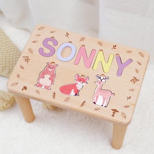 May include: A wooden step stool with the name "SONNY" spelled out in colorful letters. The stool is decorated with a bear, fox, and deer, and has a light wood finish.