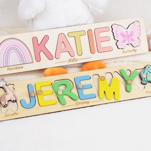 May include: Two rectangular wooden name puzzles. The top puzzle features the name KATIE in pastel letters, a rainbow, and a pink butterfly. The bottom puzzle features the name JEREMY in colorful letters with small knobs, a giraffe, and a green frog.