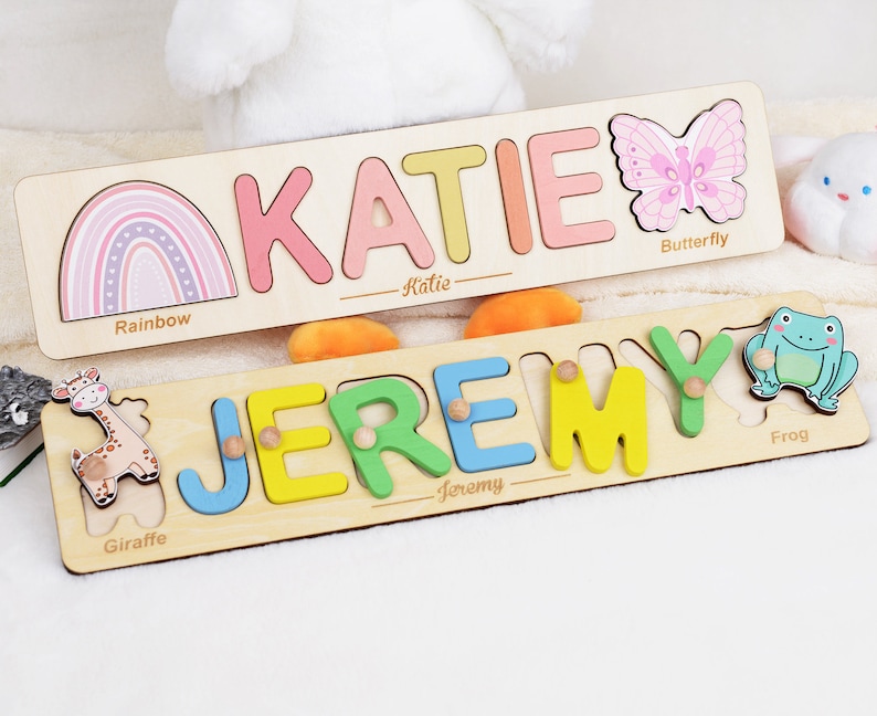 May include: Two rectangular wooden name puzzles. The top puzzle features the name KATIE in pastel letters, a rainbow, and a pink butterfly. The bottom puzzle features the name JEREMY in colorful letters with small knobs, a giraffe, and a green frog.