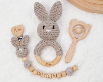 May include: A beige crocheted bunny rattle, a wooden rattle with the name "Mila" engraved on it, and a wooden teething necklace with the name "Mila" engraved on the beads.