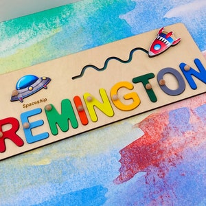 May include: A wooden name puzzle featuring the name "REMINGTON" in colourful letters. The puzzle includes a spaceship and a rocket ship.