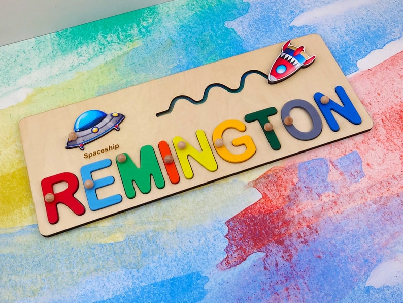 May include: A wooden name puzzle featuring the name "REMINGTON" in colourful letters. The puzzle includes a spaceship and a rocket ship.