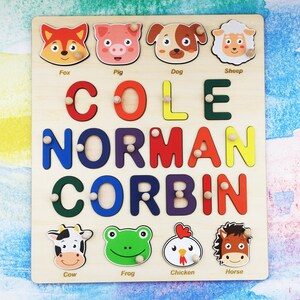 May include: A wooden puzzle with colorful letters spelling out the names "COLE NORMAN CORBIN". The puzzle has animal illustrations with the names "Fox", "Pig", "Dog", "Sheep", "Cow", "Frog", "Chicken", and "Horse" below them.