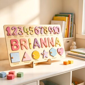 Op de afbeelding: Een houten puzzelbord met de naam BRIANNA in grote, kleurrijke letters, de cijfers &eacute;&eacute;n tot en met tien en diverse vormen zoals een vierkant, driehoek, cirkel, ster, zeshoek en hart, elk voorzien van een klein knopje om vast te pakken.