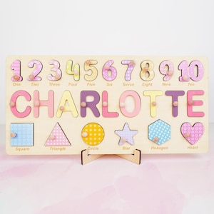 Op de afbeelding: Een rechthoekig houten puzzelbord met de naam CHARLOTTE in grote gekleurde letters, de cijfers &eacute;&eacute;n tot en met tien, en zes geometrische vormen waaronder een vierkant, driehoek, cirkel, ster, zeshoek en hart, elk met een klein knopje.
