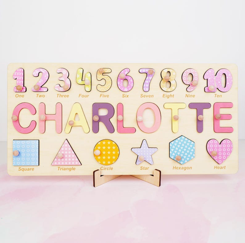 Op de afbeelding: Een rechthoekig houten puzzelbord met de naam CHARLOTTE in grote gekleurde letters, de cijfers &eacute;&eacute;n tot en met tien, en zes geometrische vormen waaronder een vierkant, driehoek, cirkel, ster, zeshoek en hart, elk met een klein knopje.