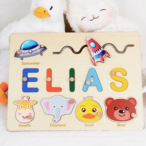 May include: Wooden personalised puzzle with the name ELIAS, featuring a spaceship, rocket, giraffe, elephant, duck, and bear. The puzzle pieces are colourful and have a cartoon style.