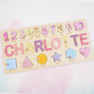 Op de afbeelding: Een houten puzzelbord met de naam CHARLOTTE in grote gekleurde letters, de cijfers &eacute;&eacute;n tot en met tien, en zes vormen waaronder een vierkant, driehoek, cirkel, ster, zeshoek en hart, elk voorzien van een kleine houten knop voor grip.
