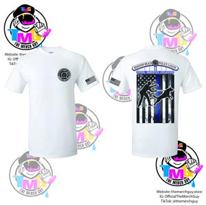 May include: White t-shirt featuring the Redondo Beach Police logo on the front and a design on the back with a police officer and canine silhouette, a blue line, and the American flag. The shirt also has a small American flag on the sleeve.