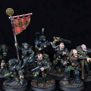 Może przedstawiać: Miniaturowe figurki jednostki wojskowej w zielonych i niebieskich mundurach, uzbrojone w broń i sztandar. Figurki znajdują się na okrągłych podstawach, z czerwonym sztandarem ze złotym emblematem.