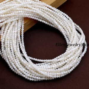 May include: Close-up of multiple strands of white, small, round beads. The beads are strung together, creating a layered effect. The beads are a creamy white color and appear to be made of a natural material. The background is a dark brown color.