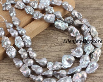 May include: A strand of grey baroque pearls, with a pearlescent sheen, arranged in three rows on a wooden surface. The pearls are irregular in shape and size, with some being more elongated than others. The pearls are strung on a white thread.