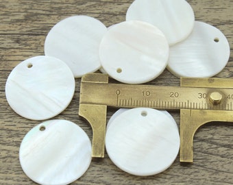 Puede incluir: Un grupo de siete discos redondos de madreperla blanca con un agujero en el centro. Los discos tienen un diámetro de aproximadamente 5 cm. Se utiliza una regla para mostrar el tamaño de los discos.