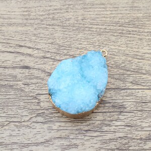 May include: A light blue crystal pendant with a gold-toned metal bail. The crystal is in a teardrop shape and has a rough, textured surface.