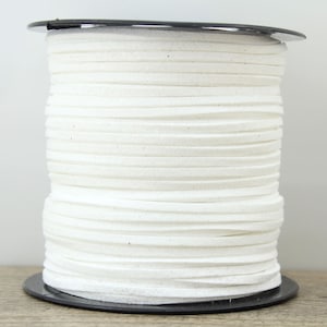 May include: A spool of off-white, flat, suede cord. The cord is tightly wound around a black plastic spool. The cord appears to be approximately 3mm wide. The background is a light neutral color.