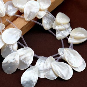 Puede incluir: Primer plano de hilos de cuentas blancas nacaradas en forma de lágrima. Las cuentas están ensartadas con un hilo fino y transparente. Las cuentas tienen una superficie lisa y brillante que refleja la luz.