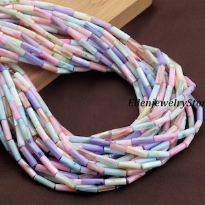 May include: A selection of pastel-coloured, rectangular beads in shades of pink, purple, blue, and cream. The beads are strung together, possibly for a necklace or bracelet. The image is set against a wooden surface and a brown background.