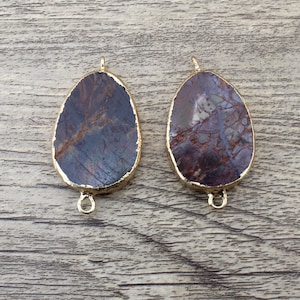 Könnte beinhalten: Zwei ovale Edelstein-Anhänger mit goldfarbenen Metallakzenten. Die Edelsteine sind braun und weiß marmoriert mit einem dunklen violetten Farbton.