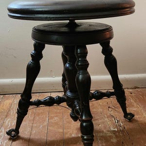 Puede incluir: Taburete de piano antiguo de color marrón oscuro con asiento redondo y tres patas curvas. El taburete tiene un reposapiés y pequeñas ruedas en los pies. La madera tiene un acabado brillante.
