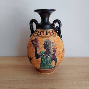 May include: A small, black and orange ceramic vase with two handles. The vase is decorated with a black silhouette of a woman holding a bird, against a yellow background.
