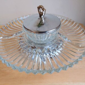 May include: A clear glass serving dish with a fluted edge and a smaller, lidded bowl in the center. The bowl has a silver lid with a gold knob.