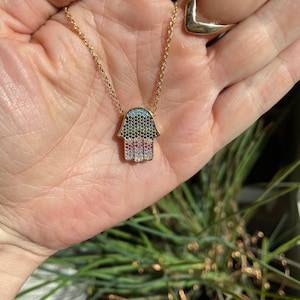 Puede incluir: Un collar de cadena de oro con un colgante de mano de hamsa. La mano de hamsa está cubierta de piedras de circonita cúbica de colores.