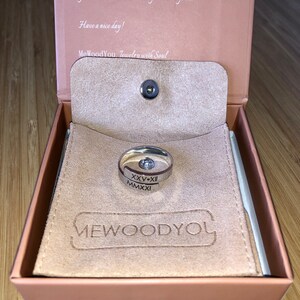 Può includere: Un anello d'argento con incisi i numeri romani XXV-XII e MMXXI. L'anello è in una scatola di camoscio marrone con le parole "MeWoodYou. Jewelry with Soul" stampate all'interno.