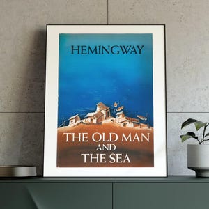 May include: A framed poster of Hemingway's "The Old Man and the Sea" with a blue and brown color scheme. The poster is displayed on a dark green cabinet with gold legs, alongside a small plant, a lamp, and decorative items.