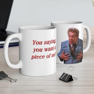 May include: White ceramic coffee mug with the text "You saying you want a piece of me?" in red. The other side of the mug features a color image of a man in a blue suit. The mug is on a light-colored surface.