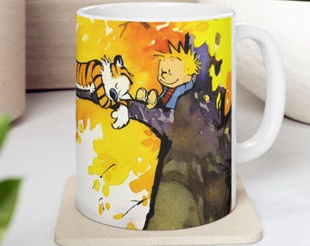 Puede incluir: Una taza de cerámica blanca con una ilustración colorida de un niño y un tigre durmiendo en un árbol. El fondo es una acuarela amarilla y naranja.