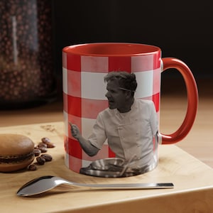May include: A red and white checkered coffee mug with a red handle and interior. The mug features a black and white image of a chef pointing. A spoon and a macaron are in the foreground.