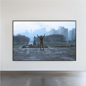 May include: A framed print of a man with arms raised in triumph, standing on a stone plaza. The background features a cityscape with tall buildings and a statue. The image is set against a hazy, overcast sky.