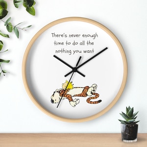 Può includere: Un orologio da parete rotondo con cornice in legno chiaro. Il quadrante bianco presenta lancette nere e il testo "There's never enough time to do all the nothing you want." Sotto c'è un cartone animato di un ragazzo e una tigre.