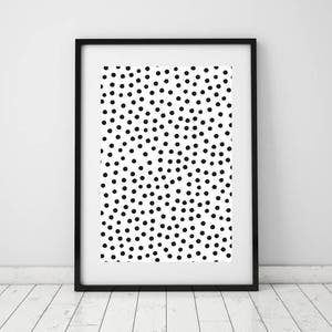 May include: Black framed art print featuring a white background with a pattern of scattered black dots. The artwork is displayed in a black frame and is leaning against a white wall and a wooden floor.