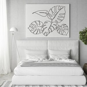 May include: A white bed with white bedding and a white headboard. A black and white line drawing of tropical leaves hangs above the bed. A small potted tree sits on the floor to the right of the bed.