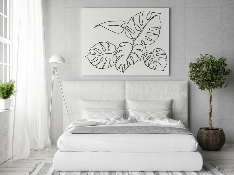 May include: A white bed with white bedding and a white headboard. A black and white line drawing of tropical leaves hangs above the bed. A small potted tree sits on the floor to the right of the bed.