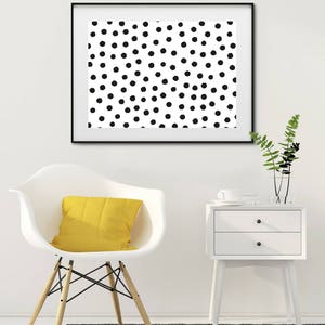 May include: A framed art print featuring a pattern of black polka dots on a white background. The artwork hangs above a white chair with a yellow cushion, a white nightstand, and a gray rug. A vase with greenery sits on the nightstand.