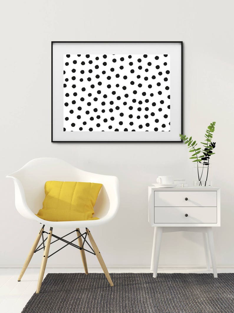 May include: A framed art print featuring a pattern of black polka dots on a white background. The artwork hangs above a white chair with a yellow cushion, a white nightstand, and a gray rug. A vase with greenery sits on the nightstand.
