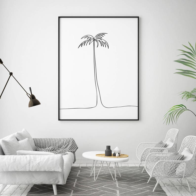 May include: A black and white line drawing of a palm tree in a black frame hanging on a white wall. The frame is above a white couch with gray throw pillows and a white coffee table with a vase and other decorative items.
