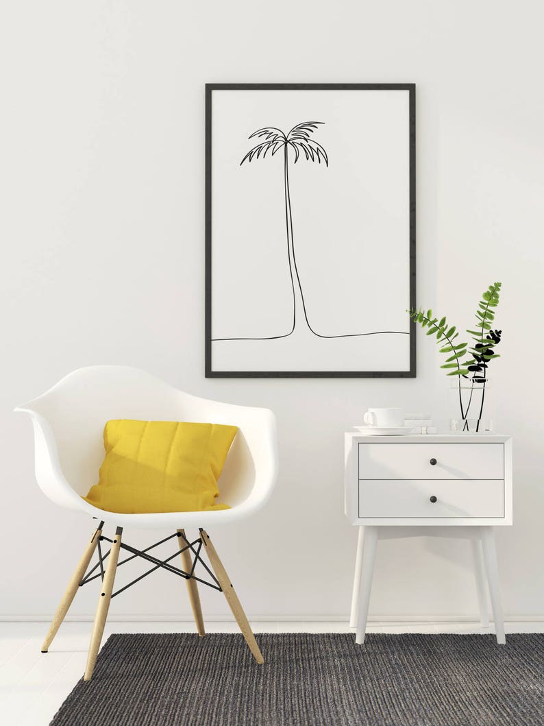 May include: A white chair with a yellow pillow sits on a gray rug in front of a white wall. A black framed print of a palm tree hangs on the wall. A white nightstand with two drawers sits next to the chair.
