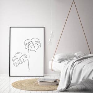 May include: A black framed print of a line drawing of two monstera leaves on a white background. The print is hanging on a white wall above a white bed with a woven rug in front of it.