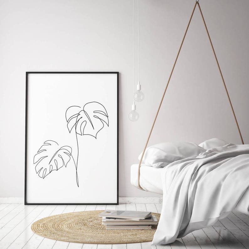 May include: A black framed print of a line drawing of two monstera leaves on a white background. The print is hanging on a white wall above a white bed with a woven rug in front of it.