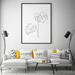 May include: A white couch with black and white patterned throw pillows and yellow throw pillows with the words "enjoy" and "life" printed on them. There is a black coffee table in front of the couch and two black floor lamps on either side of the couch. A framed black and white line drawing of two monstera leaves hangs on the wall behind the couch.
