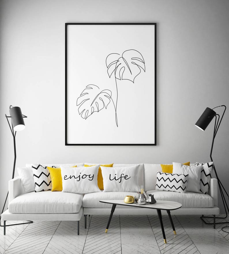 May include: A white couch with black and white patterned throw pillows and yellow throw pillows with the words "enjoy" and "life" printed on them. There is a black coffee table in front of the couch and two black floor lamps on either side of the couch. A framed black and white line drawing of two monstera leaves hangs on the wall behind the couch.