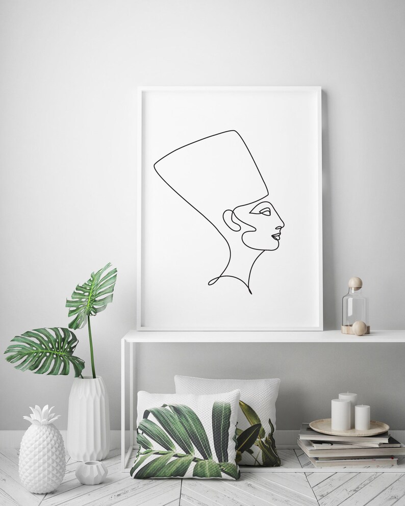 May include: A black and white line drawing of a woman wearing a headdress, printed on a white poster and framed in a white frame. The poster is hanging on a white wall above a white shelf with a white vase, a pineapple, and a glass bottle.