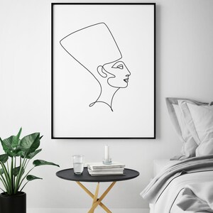 May include: Black and white line art print of a woman wearing a headdress. The print is framed in a black frame and hanging on a white wall above a black side table with a glass of water and books on it.