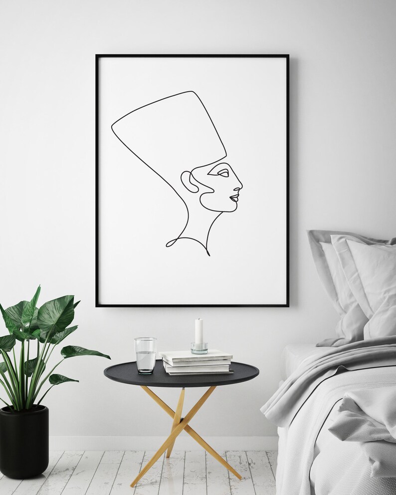 May include: Black and white line art print of a woman wearing a headdress. The print is framed in a black frame and hanging on a white wall above a black side table with a glass of water and books on it.